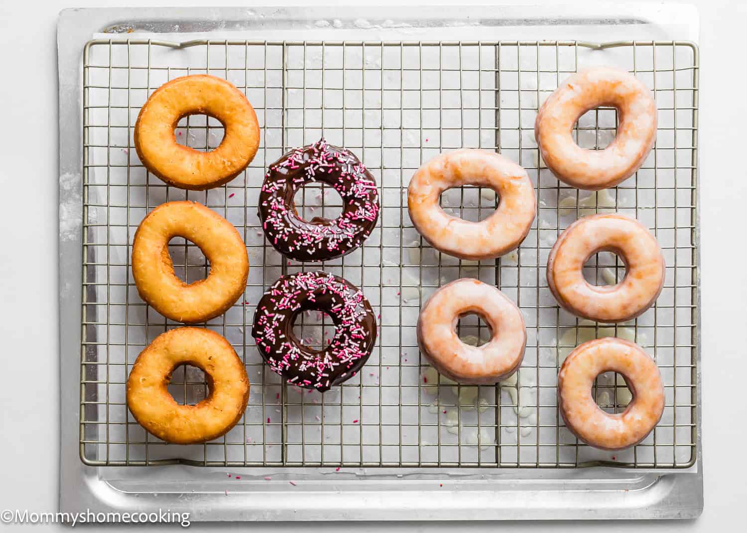 A cooling rack holds an assortment of treats: three plain donuts, two chocolate donuts with pink sprinkles, and six glazed donuts. Perfect for those who love quick and easy indulgence, these best eggless cake donuts are truly delightful on a metal tray.