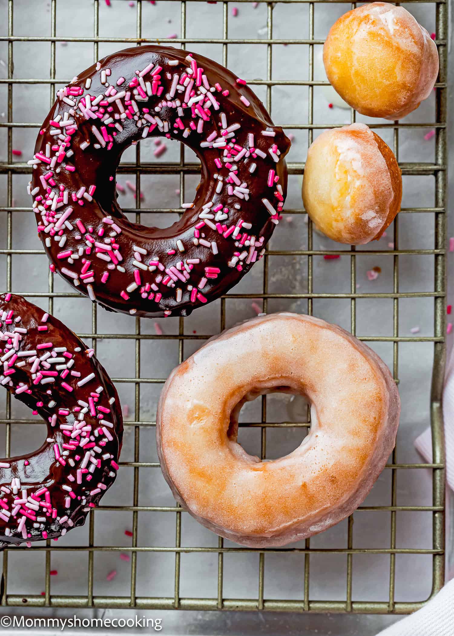 Two chocolate-glazed donuts with sprinkles and one plain-glazed donut rest on a cooling rack, accompanied by two small donut holes. These treats are reminiscent of the best eggless cake donuts—quick and easy delights for any sweet tooth!.