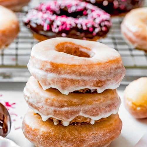 A stack of three glazed donuts, reminiscent of the best eggless cake donuts—quick and easy to make. In the background, more donuts, some adorned with pink sprinkles, rest on a wire rack.