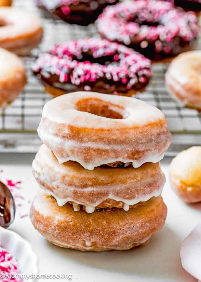 A stack of three glazed donuts, reminiscent of the best eggless cake donuts—quick and easy to make. In the background, more donuts, some adorned with pink sprinkles, rest on a wire rack.