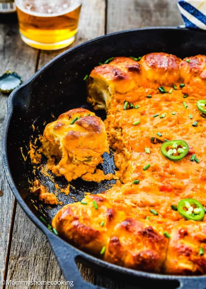 A skillet of Cheesy Taco Dip is topped with sliced jalapeños and chives, surrounded by warm garlic pretzel dippers on a wooden table. A glass of beer is partially visible in the background, promising a perfect pairing for the flavorful snack.