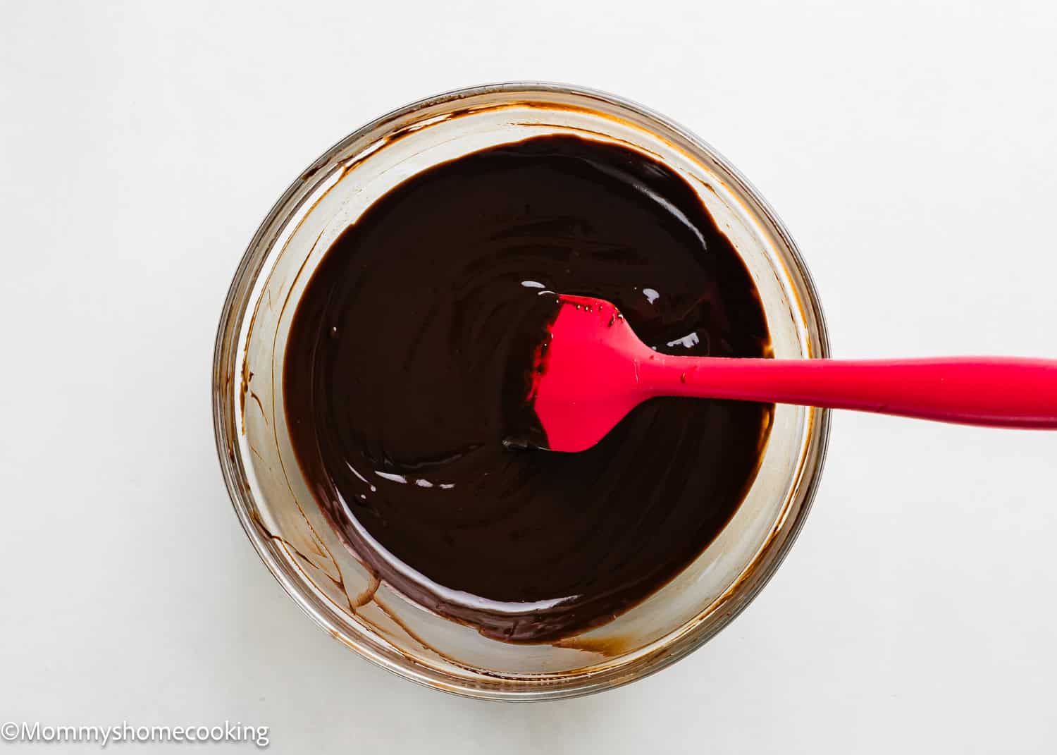 A glass bowl filled with smooth melted chocolate being stirred by a red spatula on a white surface, perfect for creating chocolate frosted cake donuts (egg-free).