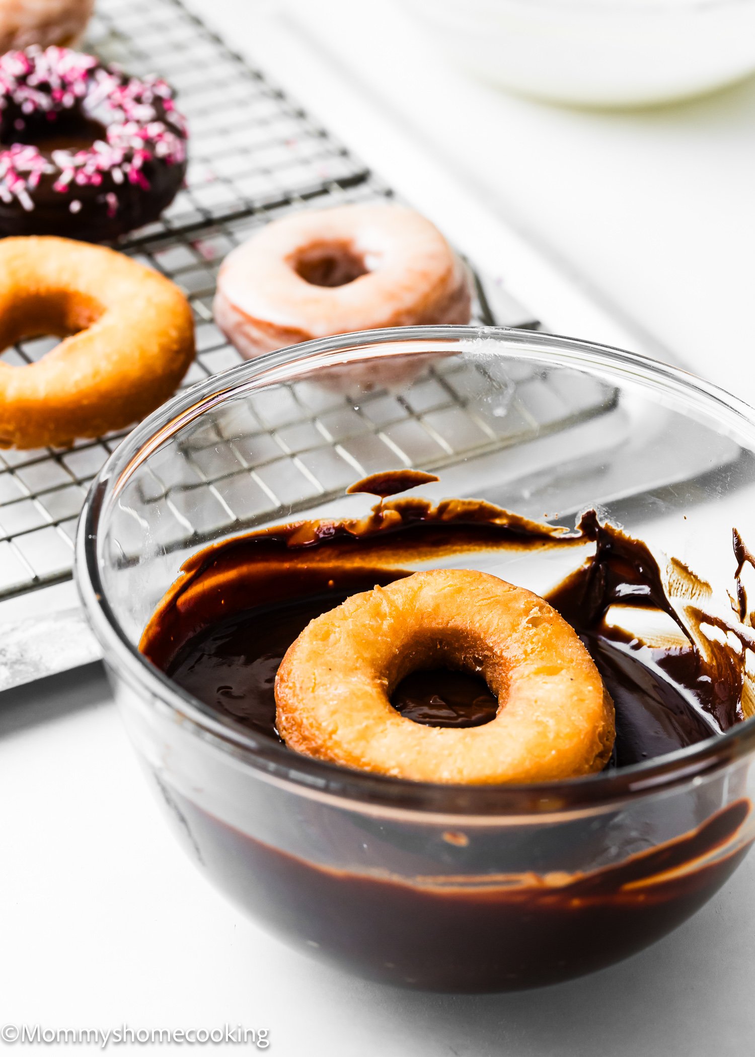 A donut is being dipped into a bowl of chocolate glaze, with chocolate frosted cake donuts (egg-free) resting on a cooling rack in the background.