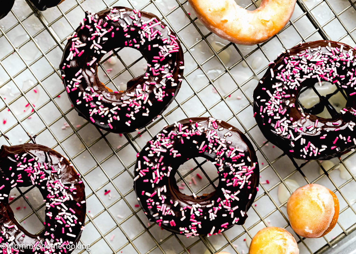 Chocolate frosted cake donuts, egg-free, with pink and white sprinkles rest on a cooling rack.