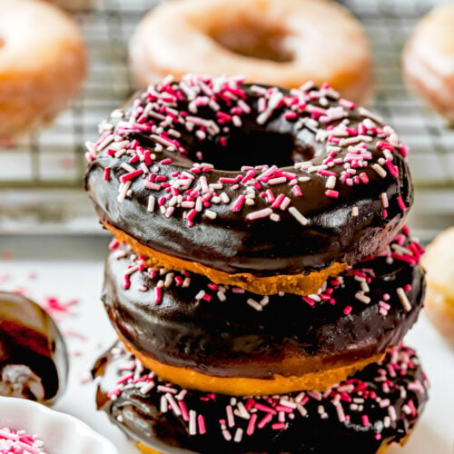 A stack of three chocolate-frosted cake donuts sits in front of a cooling rack lined with plain ones, each donut topped with vibrant pink and white sprinkles.