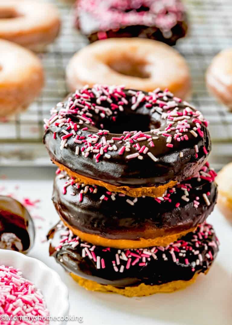 A stack of three chocolate-frosted cake donuts sits in front of a cooling rack lined with plain ones, each donut topped with vibrant pink and white sprinkles.