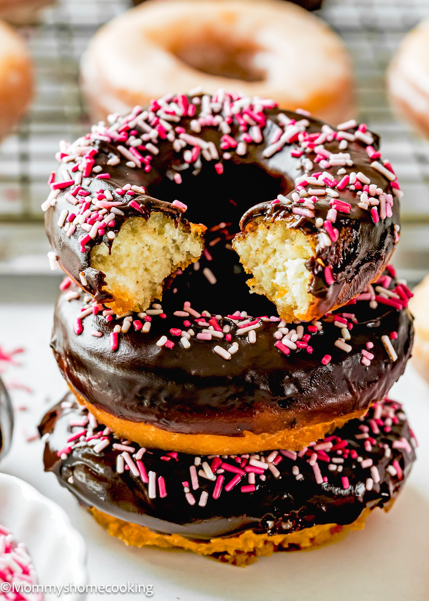 Three chocolate-frosted cake donuts, egg-free, are stacked with pink and white sprinkles; the top donut has a bite taken out of it.