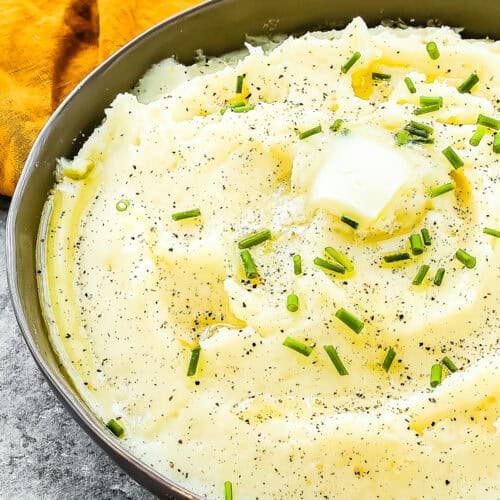 A bowl of easy, creamy mashed potatoes garnished with chives, a dollop of butter melting on top, and sprinkled with black pepper sits invitingly on a table adorned with an orange napkin. This smooth recipe ensures no lumps for the perfect side dish.