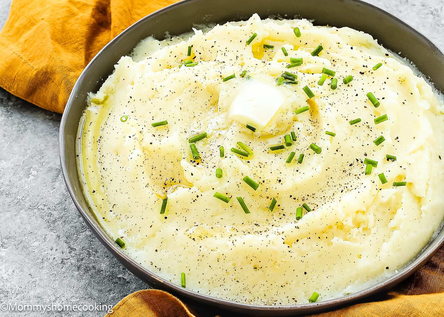 Bowl of smooth, creamy mashed potatoes—an easy, lump-free recipe—topped with a pat of butter, chopped chives, and black pepper. Placed on a gray surface next to an orange napkin for the perfect comforting side dish.