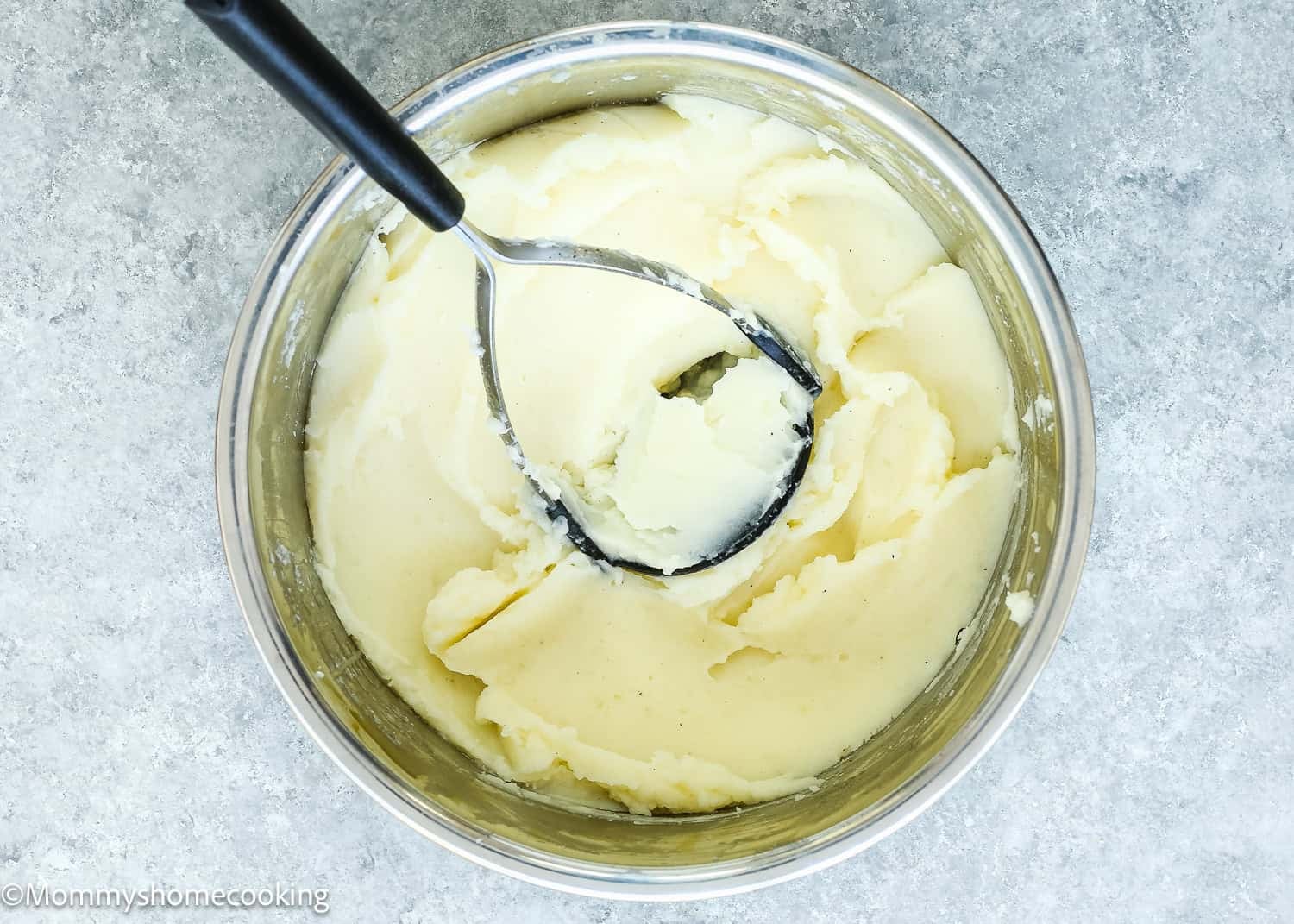 A bowl of creamy mashed potatoes sits ready to serve, crafted following an easy recipe for ultra-smooth, lump-free results. The black-handled masher rests on the gray surface, showcasing perfect preparation.