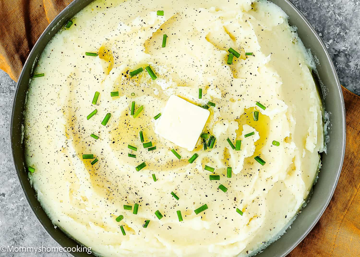 A bowl of easy creamy mashed potatoes, made smooth with no lumps, is topped with a pat of butter, chopped chives, and a sprinkle of black pepper.