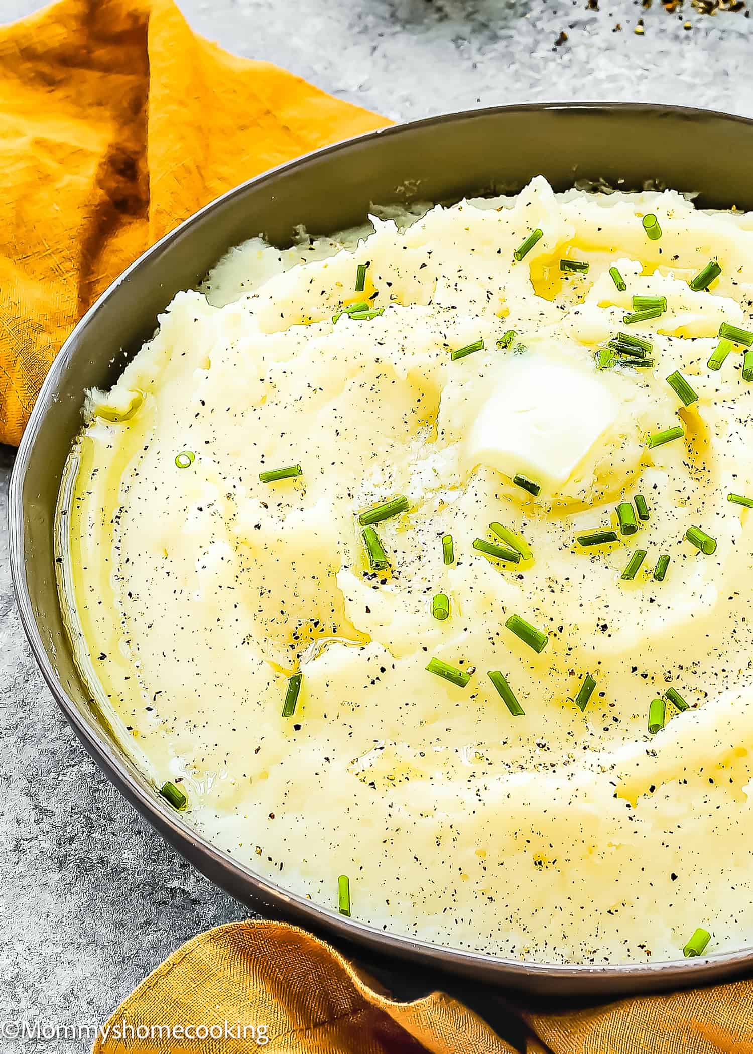 A bowl of easy, creamy mashed potatoes garnished with chives, a dollop of butter melting on top, and sprinkled with black pepper sits invitingly on a table adorned with an orange napkin. This smooth recipe ensures no lumps for the perfect side dish.