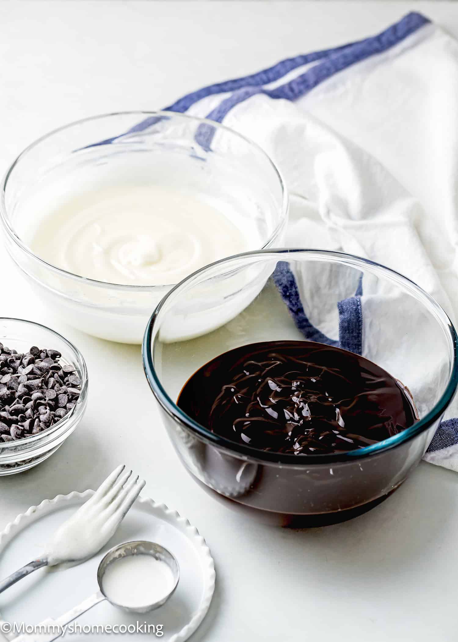 Two bowls, one filled with chocolate glaze and the other with creamy vanilla donut glaze, sit invitingly on the counter. Nearby, a small dish of chocolate chips, a fork, and a cloth complete the easy setup for crafting sweet delights.