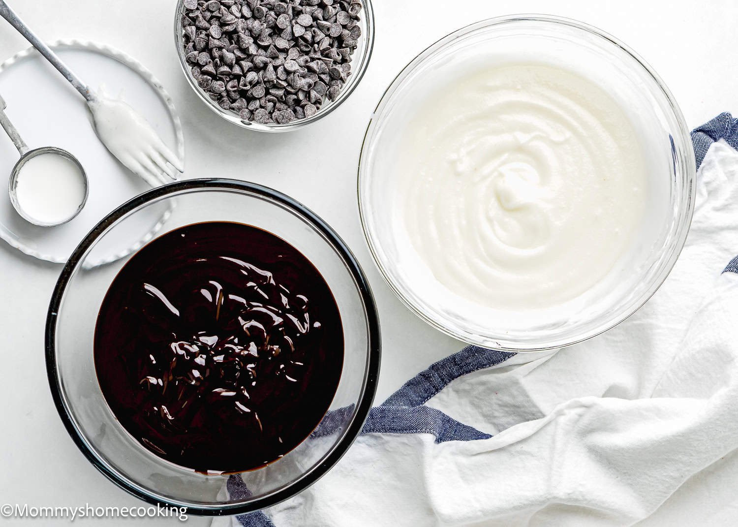 Two bowls on a white surface: one with chocolate glaze, perfect for an easy donut glaze, and one with vanilla glaze. A small bowl of chocolate chips and a teaspoon are beside them, ready for your delicious creation. A cloth napkin is nearby.