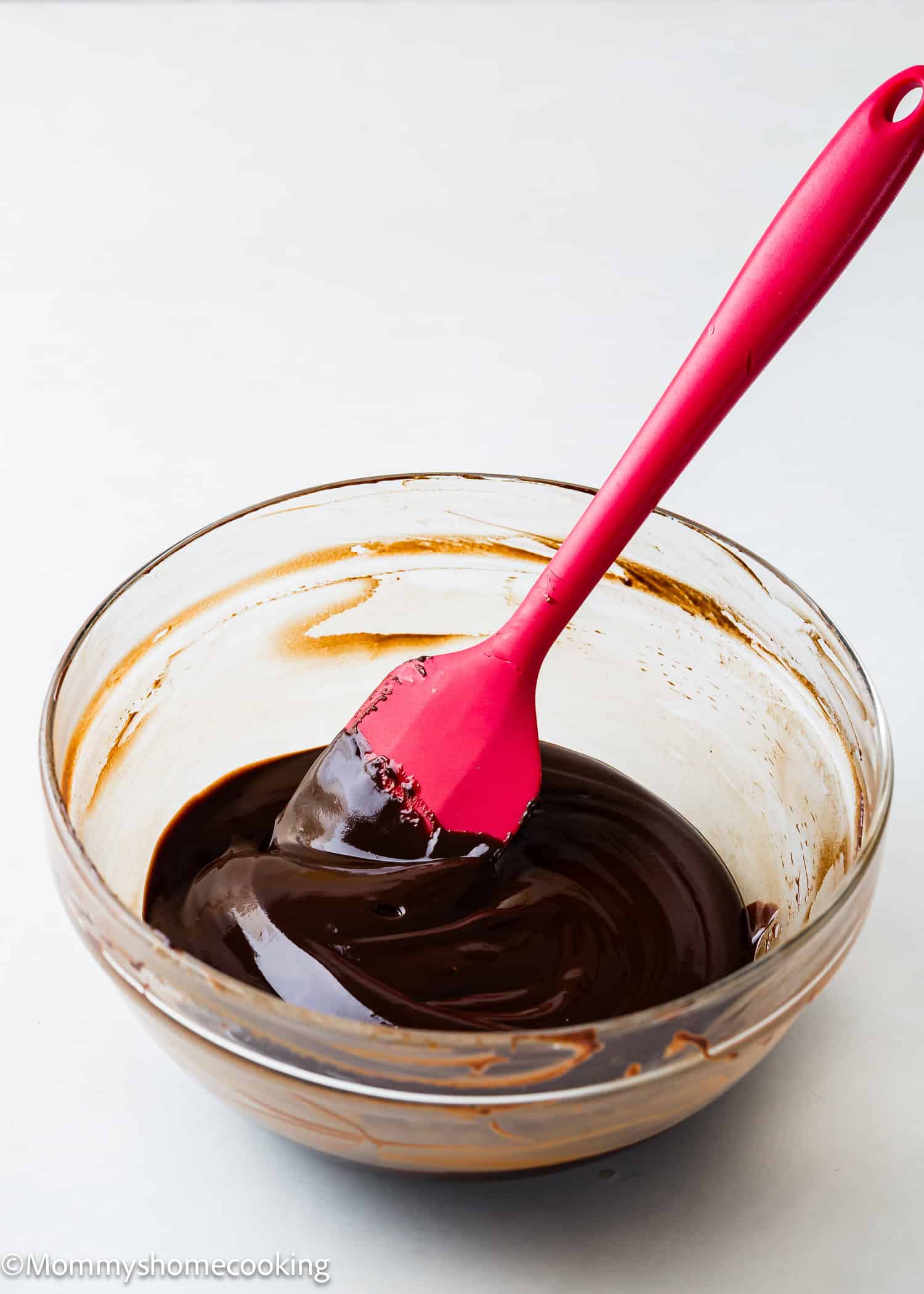 A glass bowl filled with chocolate glaze, is being stirred with a red spatula.