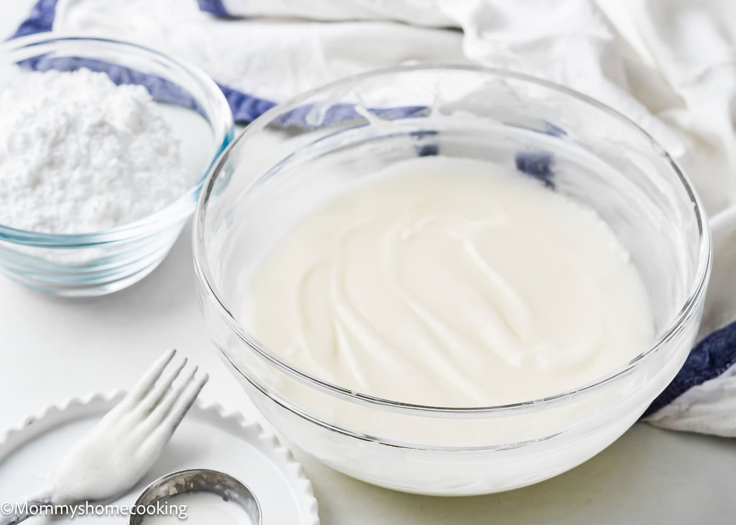 A bowl of smooth white icing, perfect for easy donut glaze, sits next to a smaller bowl of powdered sugar on a white surface with a fork and cloth nearby.