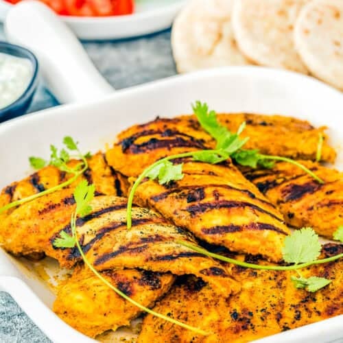Easy Chicken Shawarma: Succulent grilled chicken pieces garnished with cilantro in a white dish, accompanied by sliced tomatoes, crisp lettuce, pita bread, and a flavorful dip in the background.
