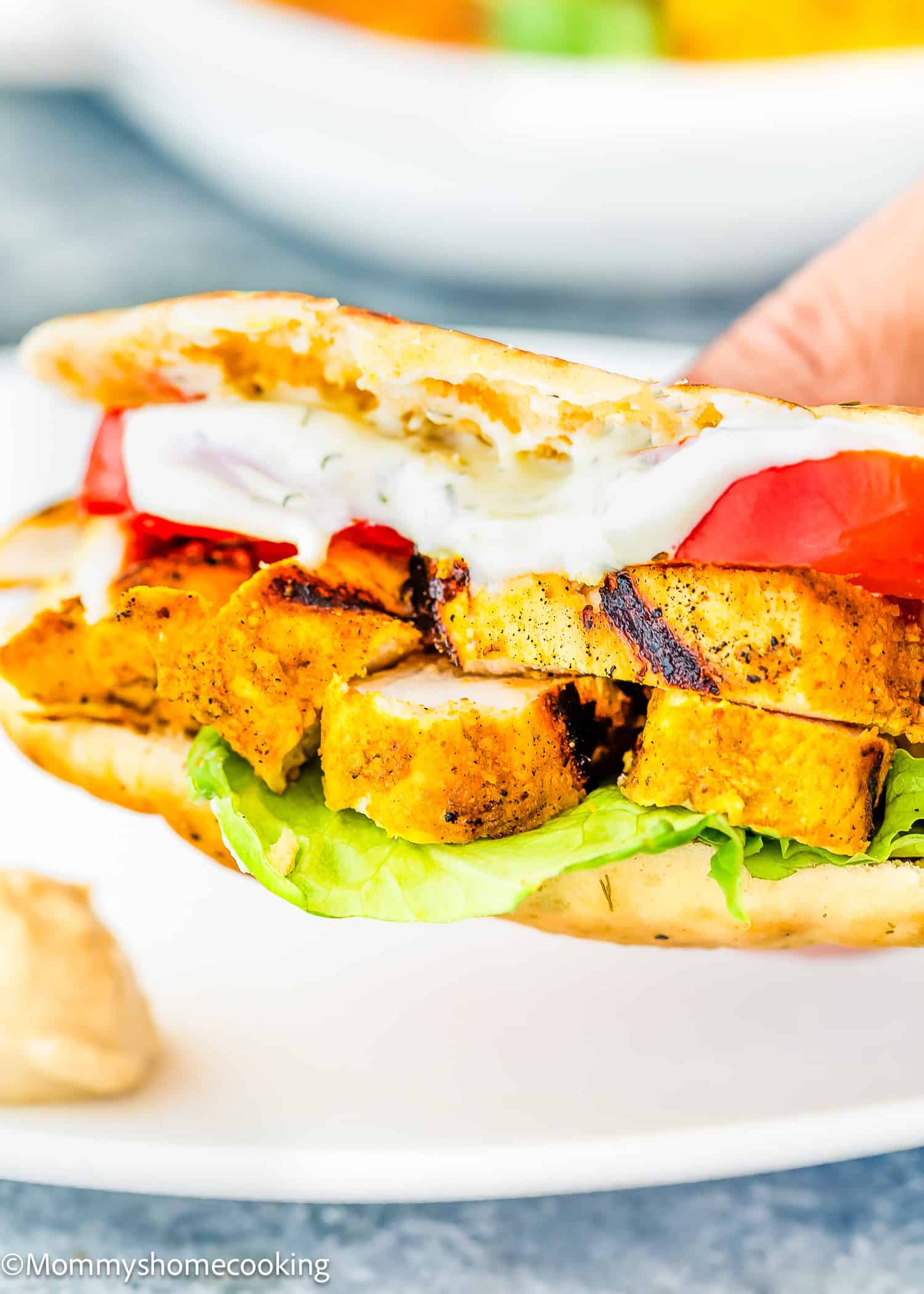 A hand holds an easy chicken shawarma in the form of a grilled chicken sandwich, complete with lettuce, tomato, and sauce nestled in soft pita bread.