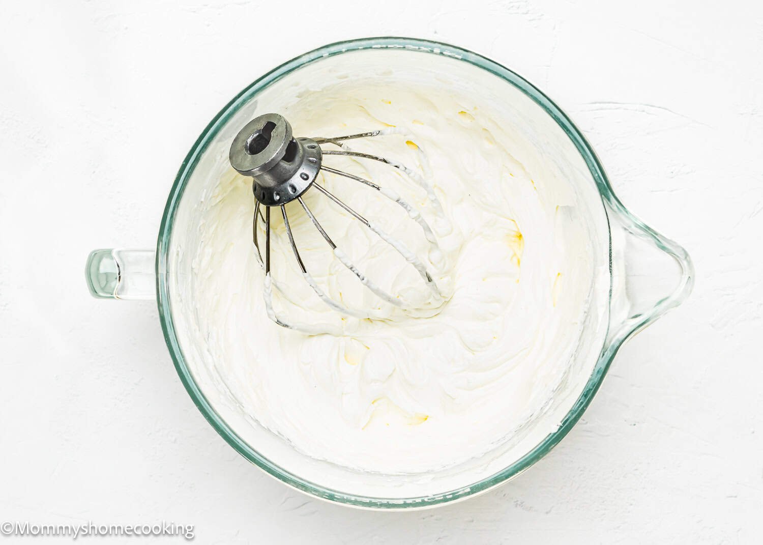 A mixing bowl brimming with easy whipped cream cheese frosting sits ready, a whisk attachment resting on top, set against a pristine white background.