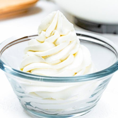 Swirled mound of Easy Whipped Cream Cheese Frosting in a clear glass bowl on a white surface.