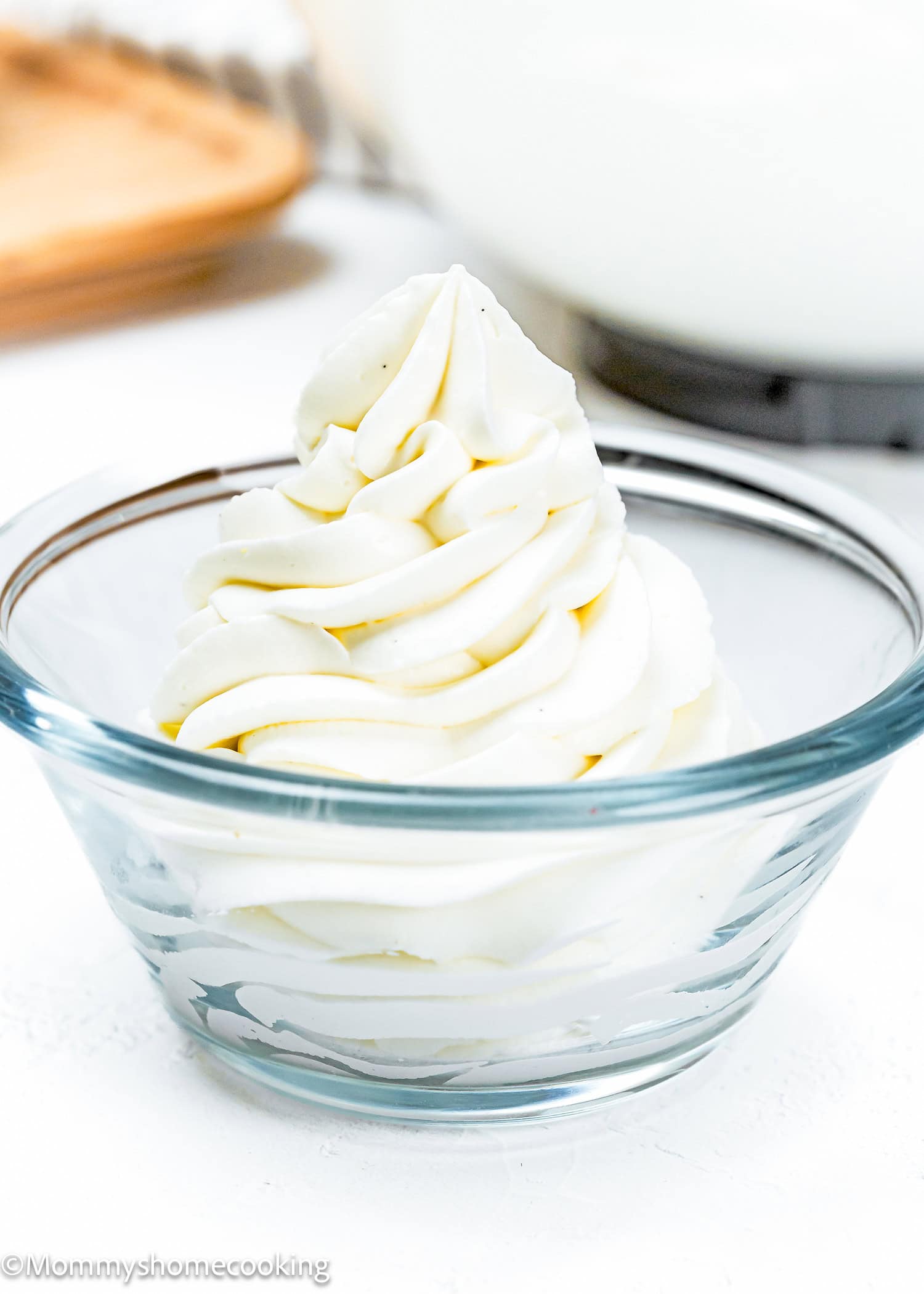 Swirled mound of Easy Whipped Cream Cheese Frosting in a clear glass bowl on a white surface.