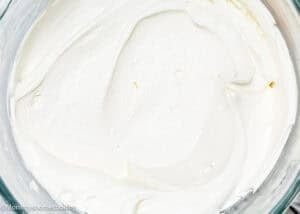 A glass bowl brimming with thick white easy whipped cream cheese frosting on a white surface, next to a wooden board with a scoop of powdered sugar and a striped cloth.