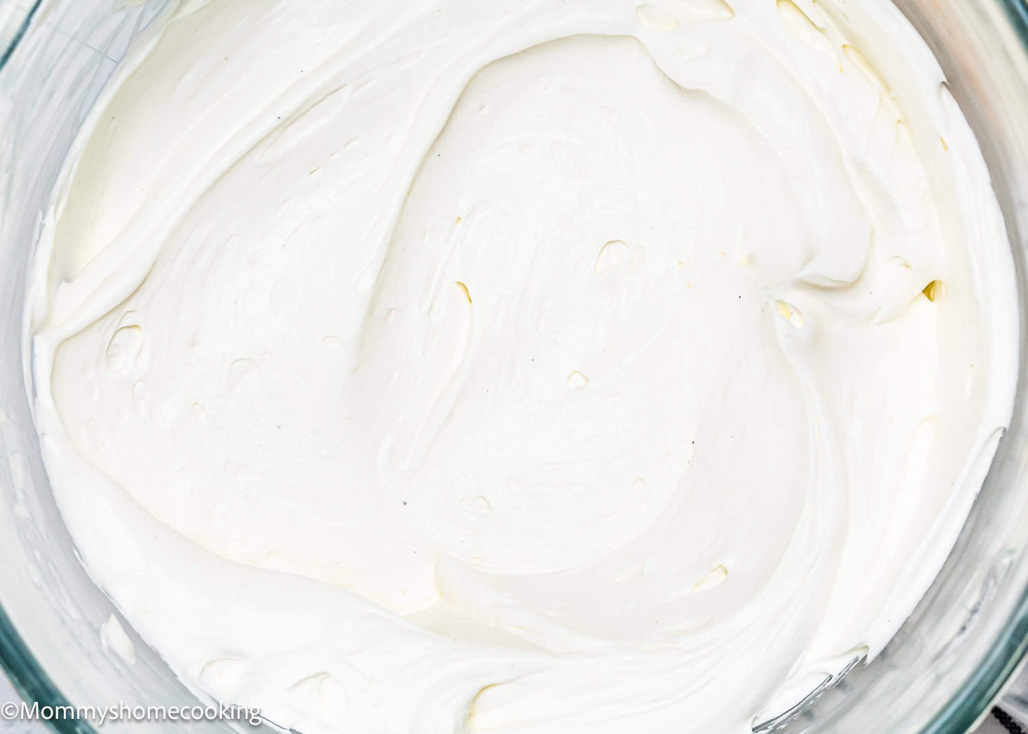 A glass bowl brimming with thick white easy whipped cream cheese frosting on a white surface, next to a wooden board with a scoop of powdered sugar and a striped cloth.
