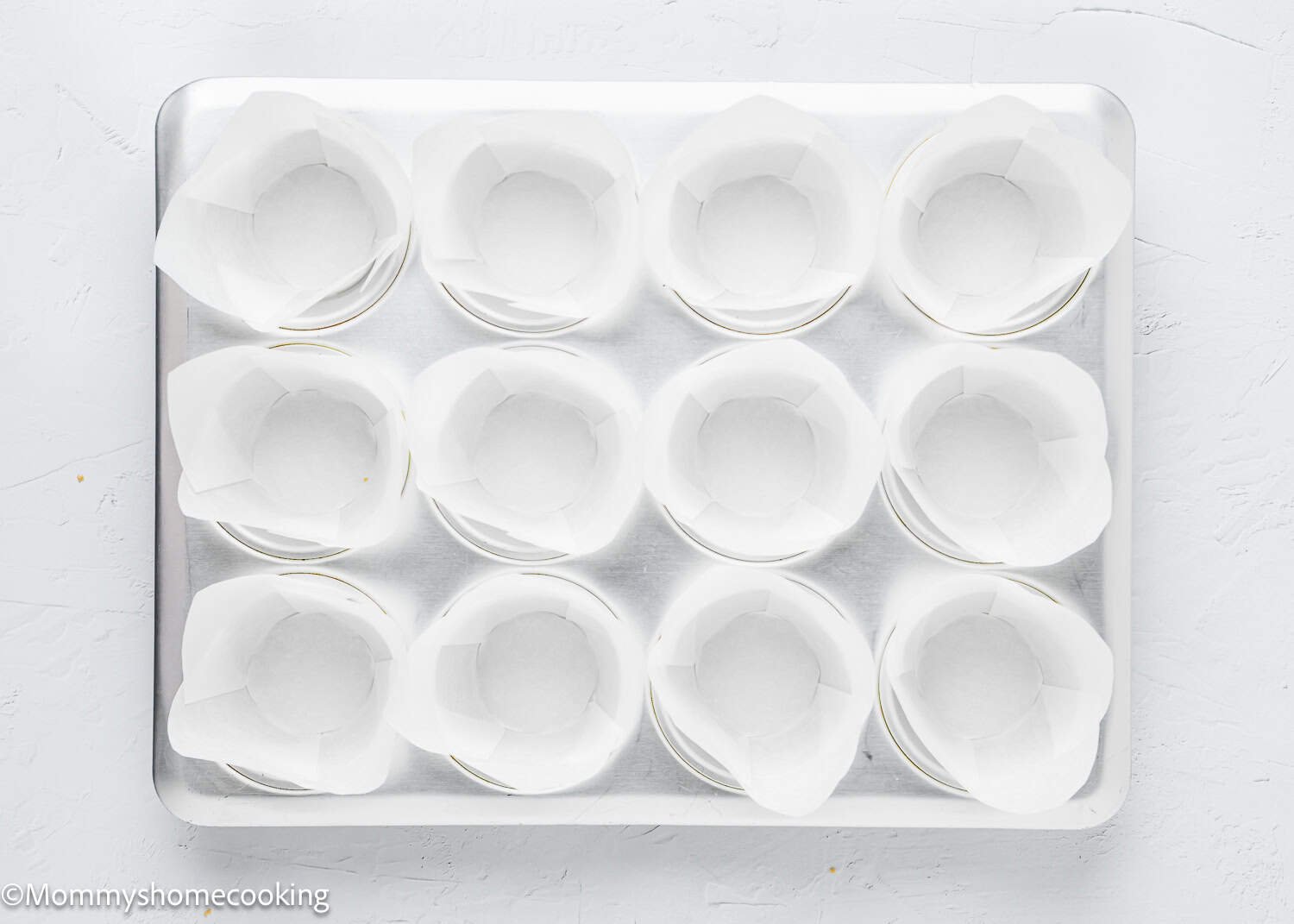 A baking tray holding twelve muffin cups lined with white parchment paper sits on a light background, ready for eggless coffee cake muffins to be baked.