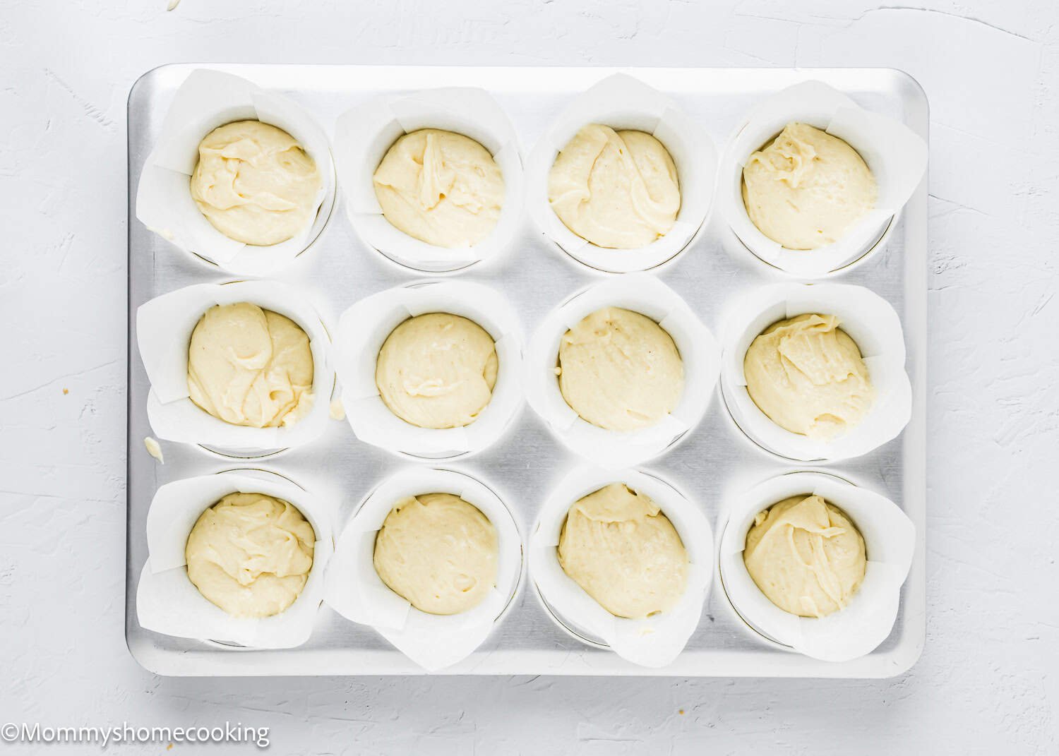Muffin tin with twelve compartments filled with raw batter for eggless coffee cake muffins in parchment liners, ready for baking on a light-colored surface.