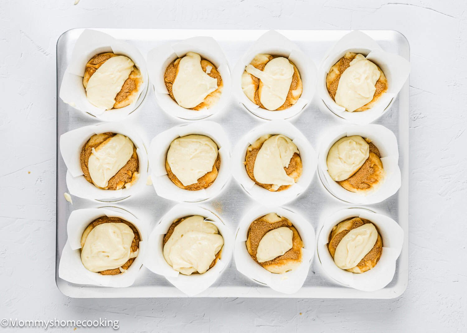 A baking tray with twelve paper liners filled with brown eggless coffee cake muffin batter, topped with dollops of cream-colored batter, ready for baking.