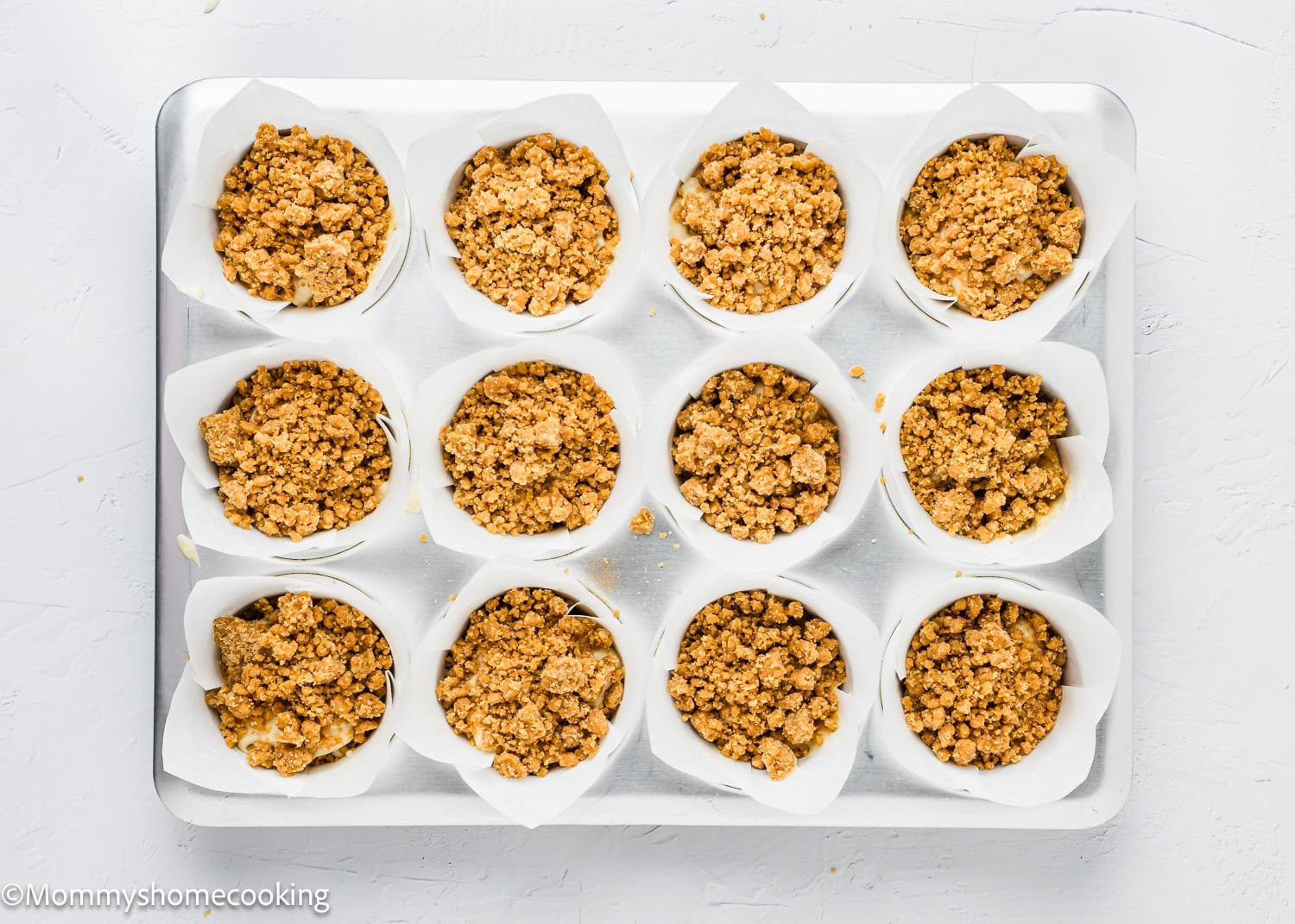 Twelve paper-lined muffin cups filled with crumbly eggless coffee cake muffin batter are arranged on a baking tray.