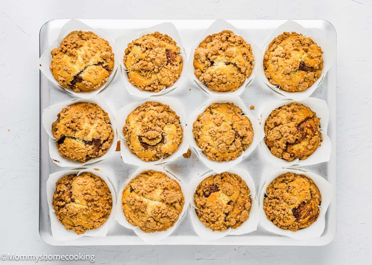 A tray of twelve eggless coffee cake muffins in white paper liners, each topped with a crumbly streusel.