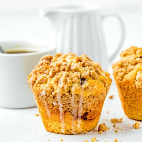 A close-up of an eggless coffee cake muffin adorned with streusel topping and glaze sits beside a white cup and a white pitcher on a light surface.