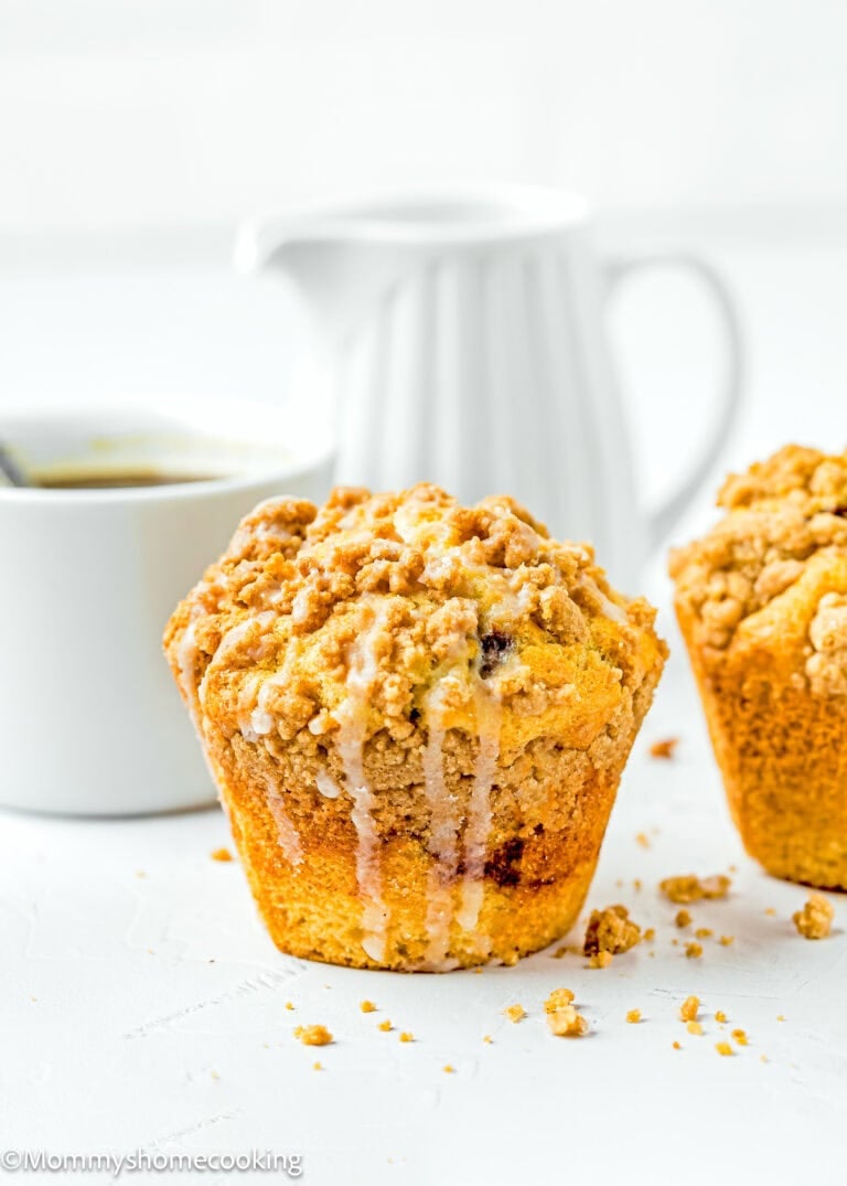 A close-up of an eggless coffee cake muffin adorned with streusel topping and glaze sits beside a white cup and a white pitcher on a light surface.