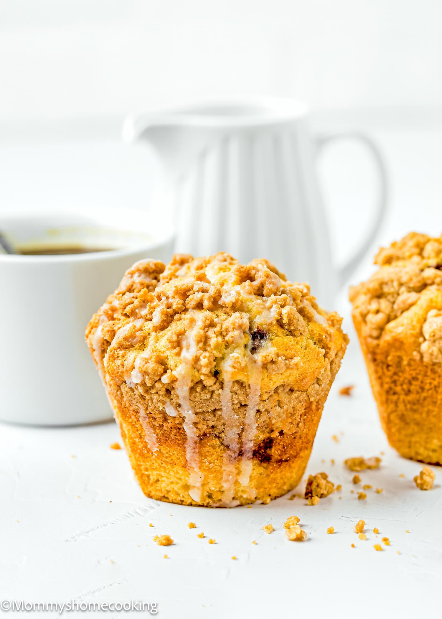 A close-up of an eggless coffee cake muffin adorned with streusel topping and glaze sits beside a white cup and a white pitcher on a light surface.