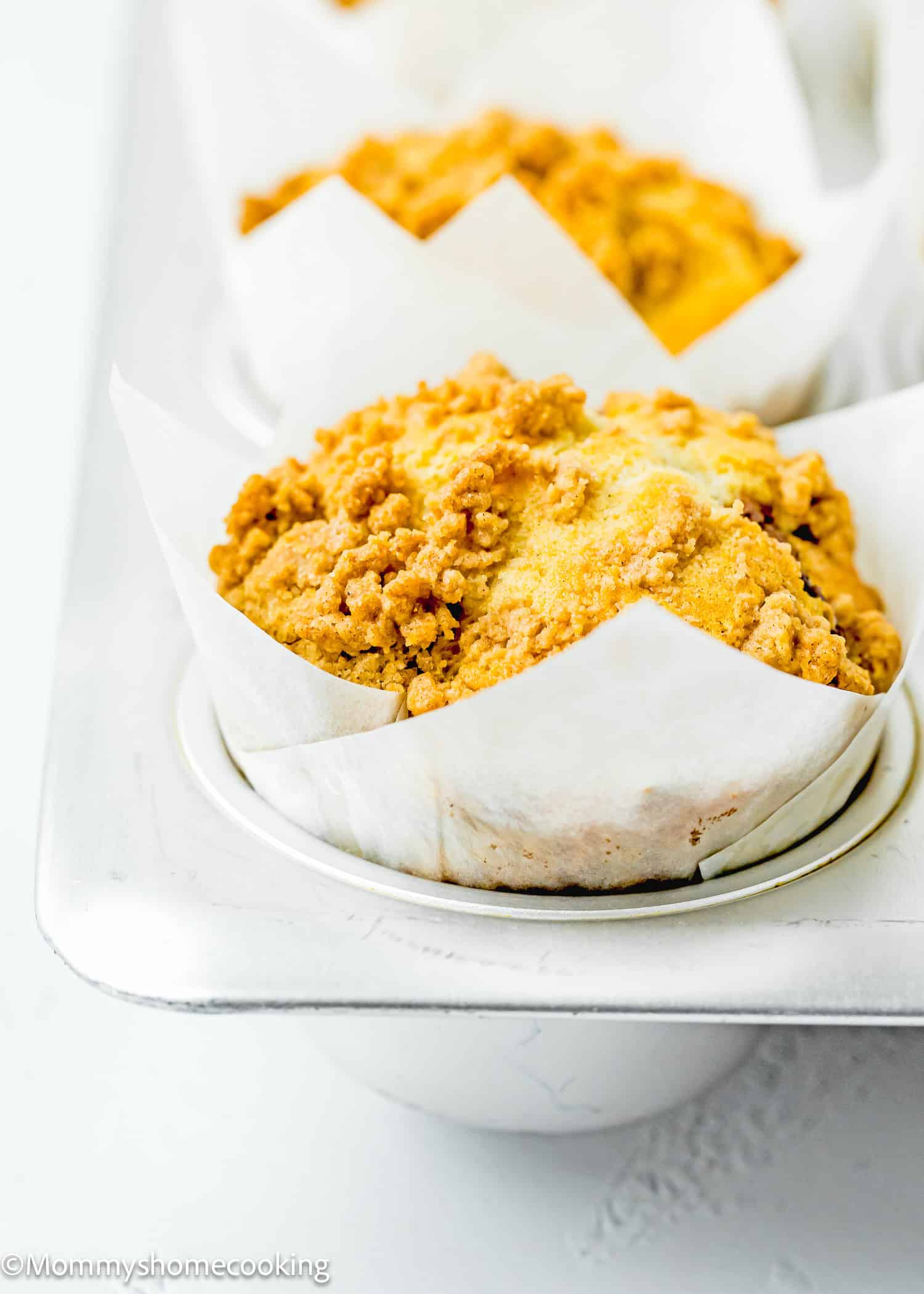 Eggless coffee cake muffin with a crumbly topping in a white paper liner, placed in a metal baking tray.
