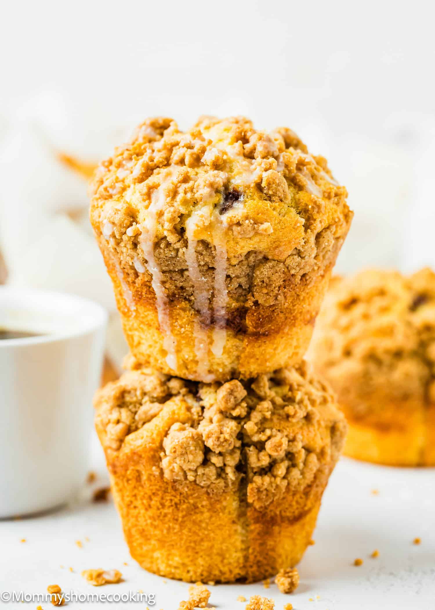Two eggless coffee cake muffins, adorned with a streusel topping, are stacked on top of each other and drizzled with glaze. In the background, a cup of coffee adds a warm touch to the scene.