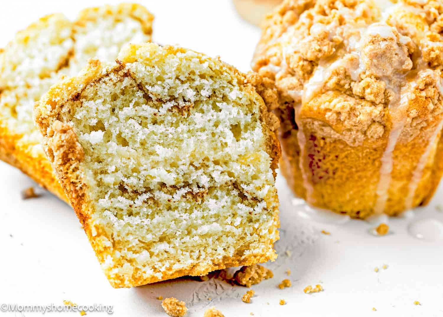 A close-up of two eggless coffee cake muffins, one whole and one halved, reveals their fluffy, crumbly texture with a streusel topping. Crumbs are artfully scattered across the white surface.