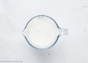A glass measuring cup filled with a creamy white liquid sits on a textured white surface, perfect for preparing eggless coffee cake muffins.