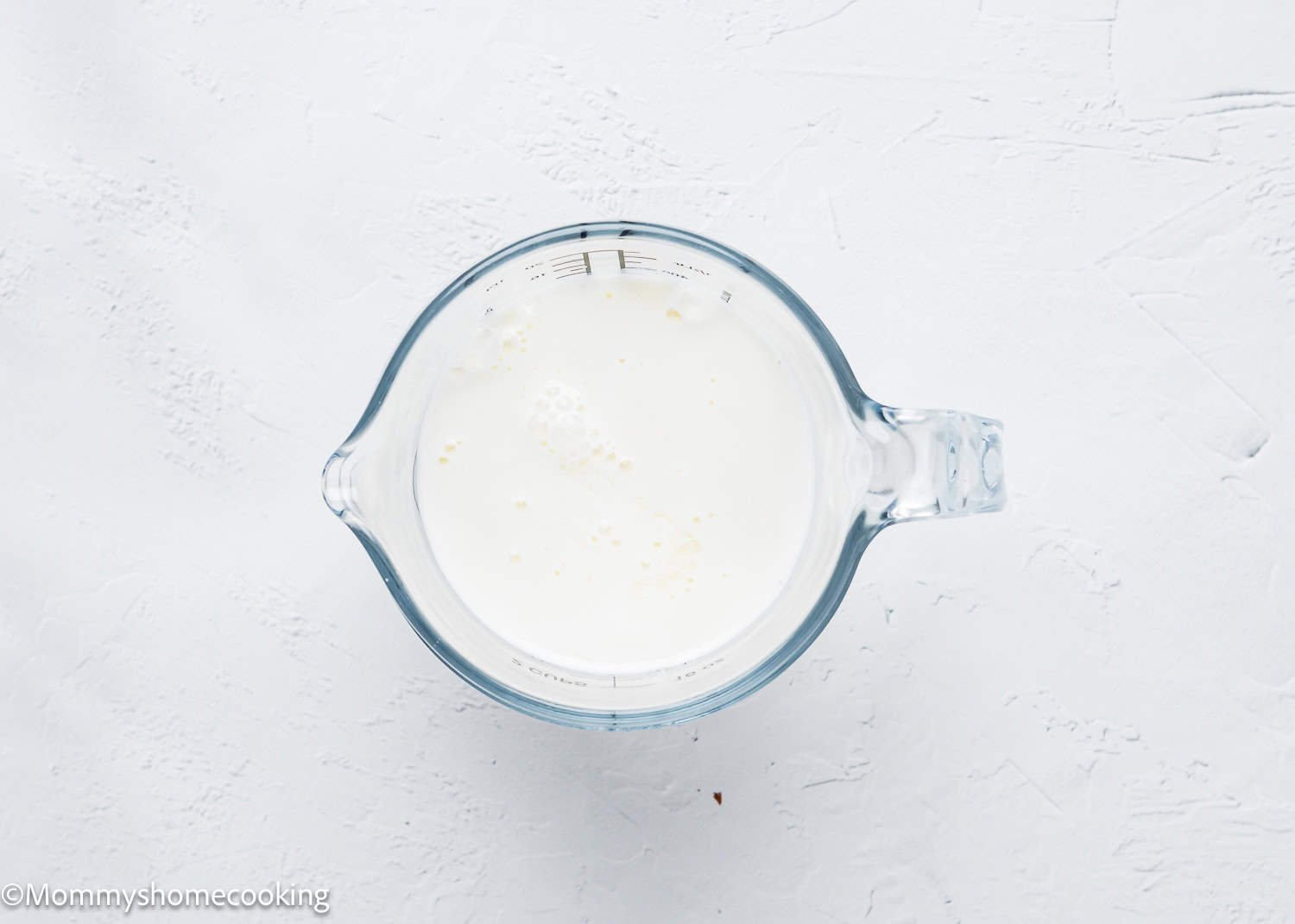 A glass measuring cup filled with a creamy white liquid sits on a textured white surface, perfect for preparing eggless coffee cake muffins.