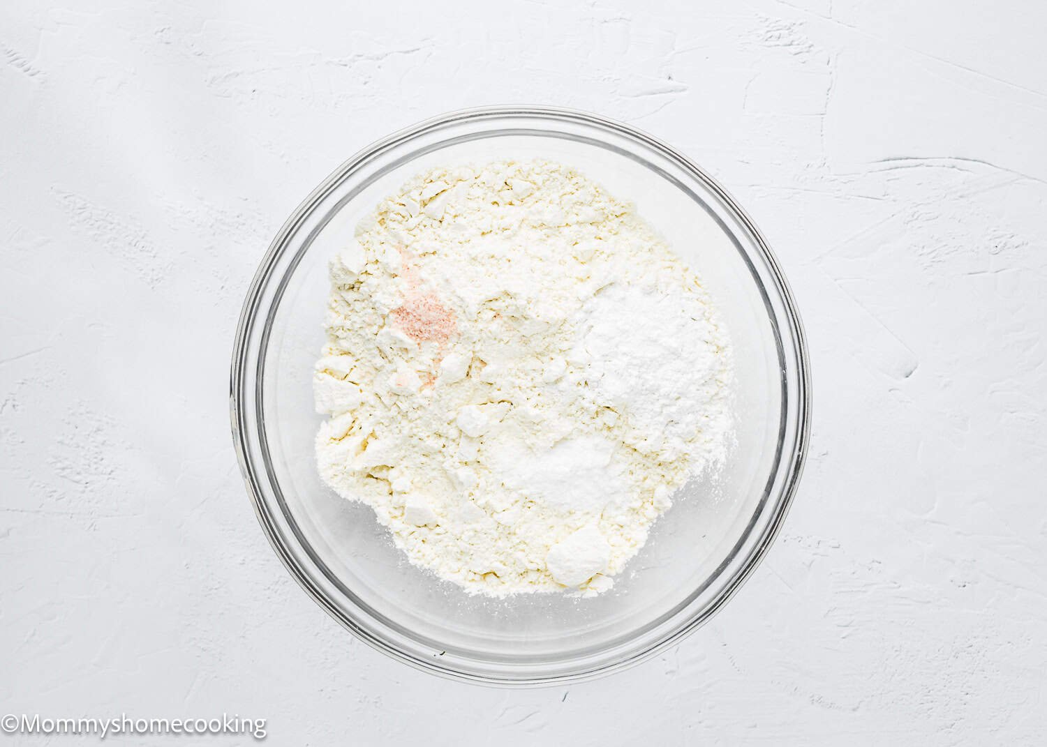 A glass bowl contains flour, baking soda, and salt on a white textured surface, ready to create delicious eggless coffee cake muffins.