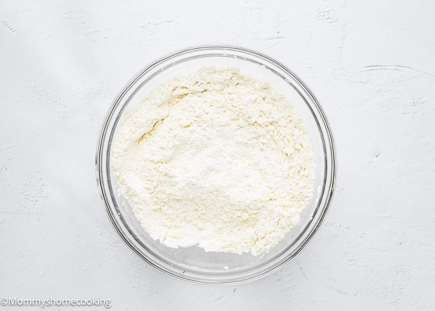 A clear glass bowl filled with white flour rests on a textured white surface, ready to craft delightful eggless coffee cake muffins.