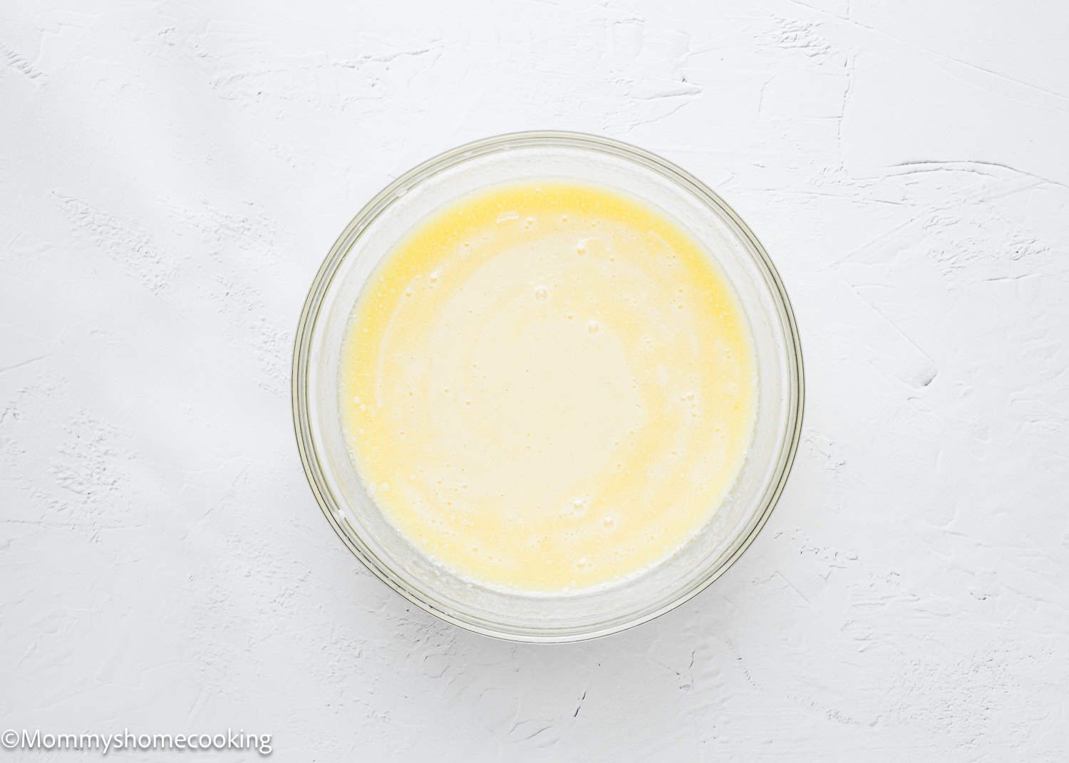 A glass bowl containing a smooth, creamy yellow mixture sits on a textured white surface, ready to be transformed into delicious eggless coffee cake muffins.
