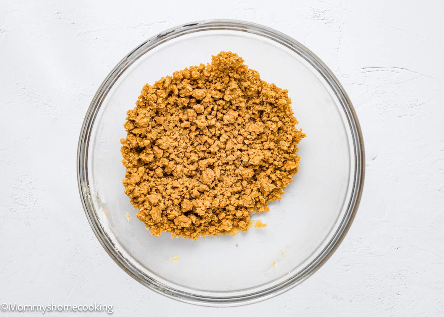 A glass bowl on a white surface holds a crumbly, brown sugar crumb perfect for making eggless coffee cake muffins.