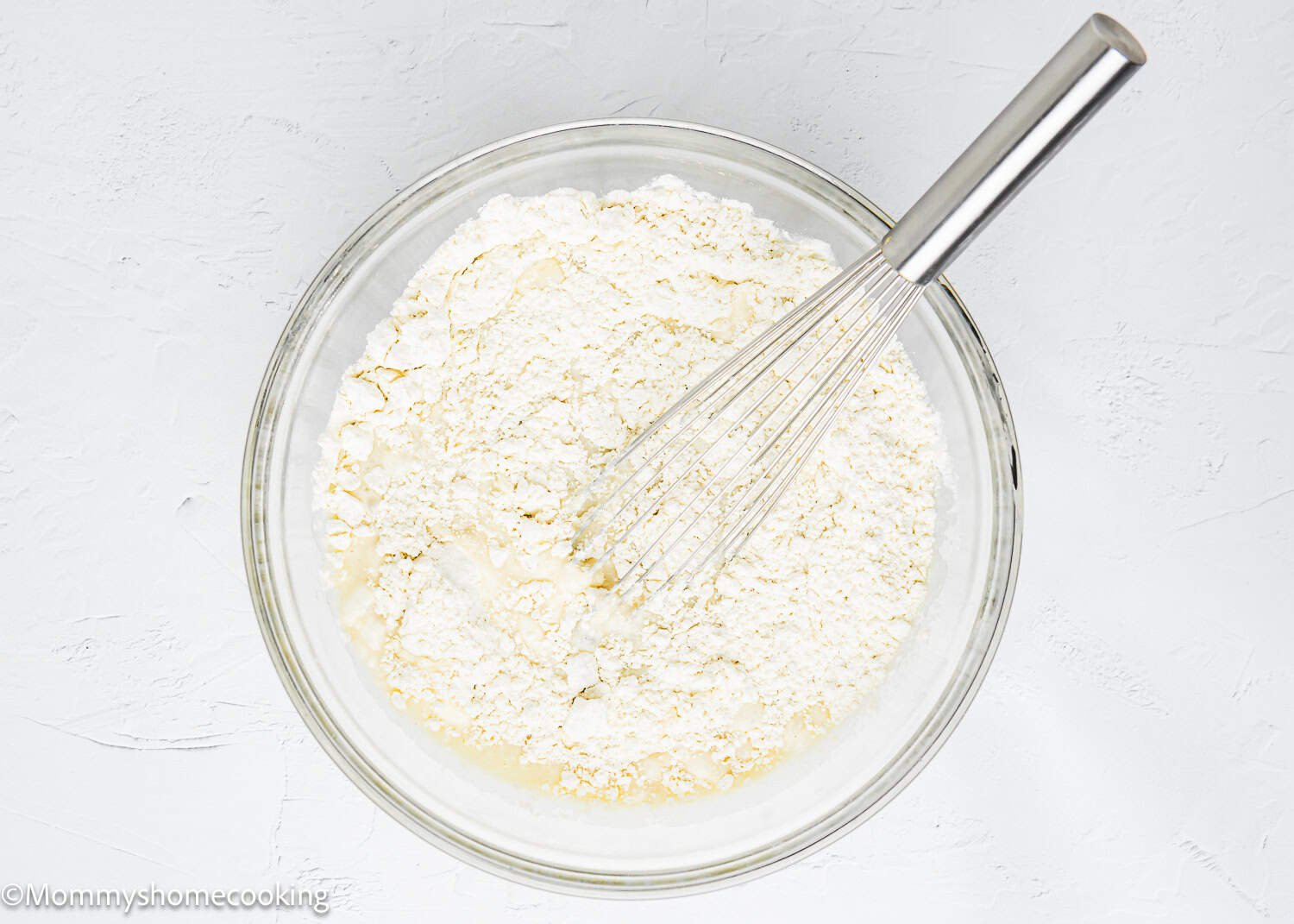 A glass bowl filled with flour and a whisk on a light background, ready for making eggless coffee cake muffins.