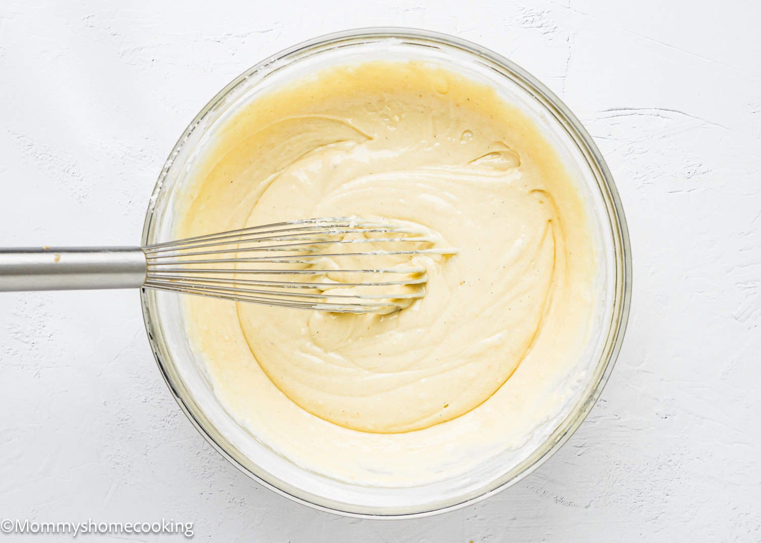 A glass bowl brimming with smooth, creamy batter for delectable eggless coffee cake muffins is being mixed with a metal whisk on a light background.
