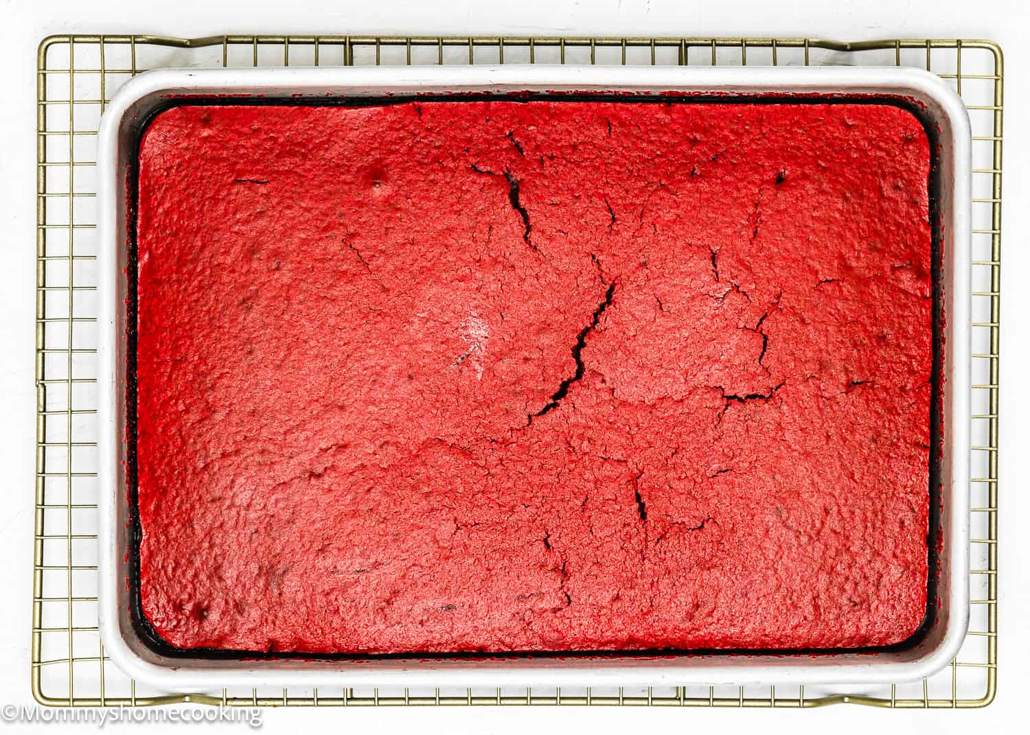 An eggless red velvet brownie poke cake with surface cracks rests in a white baking tray on a wire cooling rack, showcasing its rich, decadent texture.