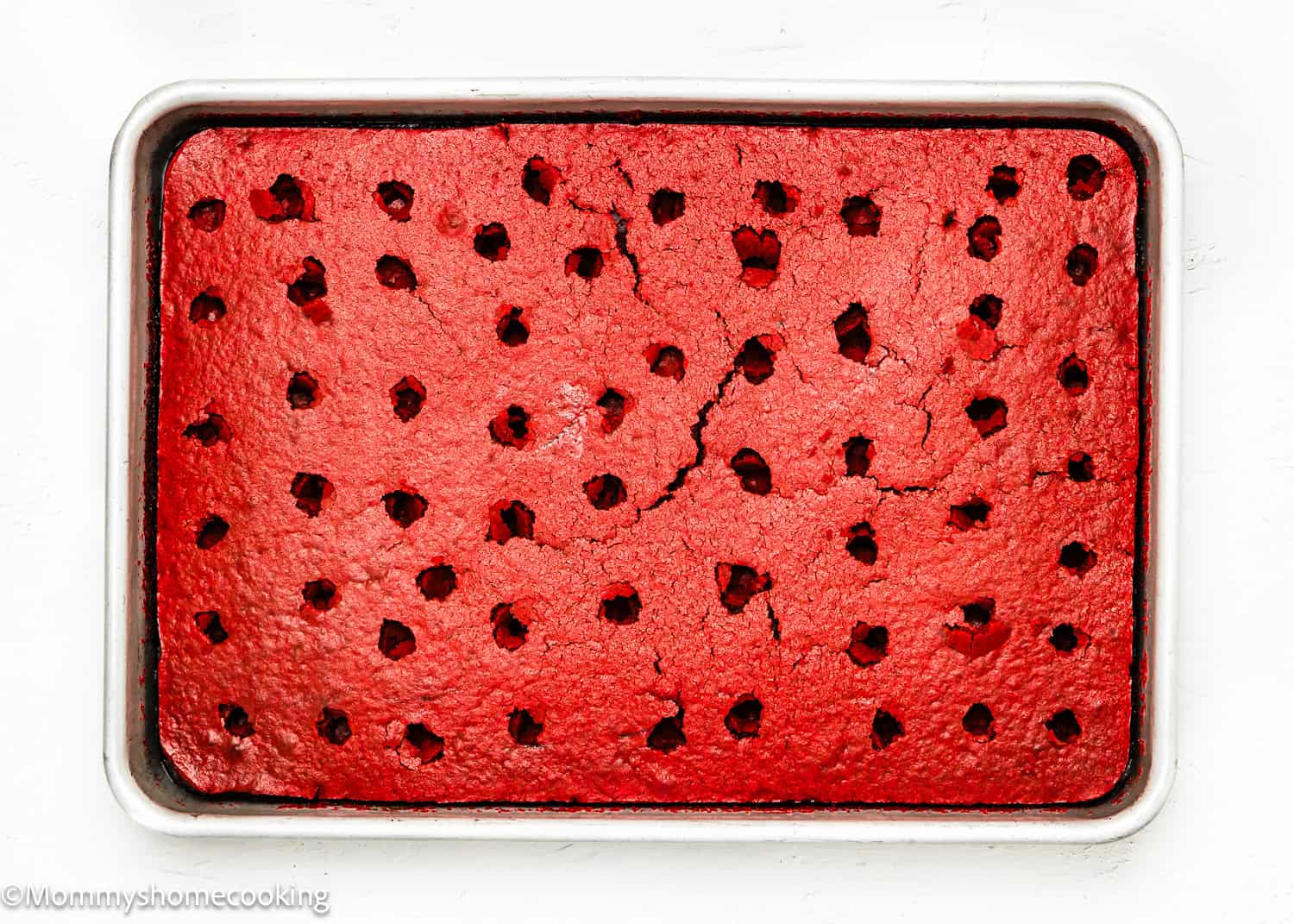 An Eggless Red Velvet Brownie Poke Cake rests in a baking pan, its surface dotted with evenly spaced holes.