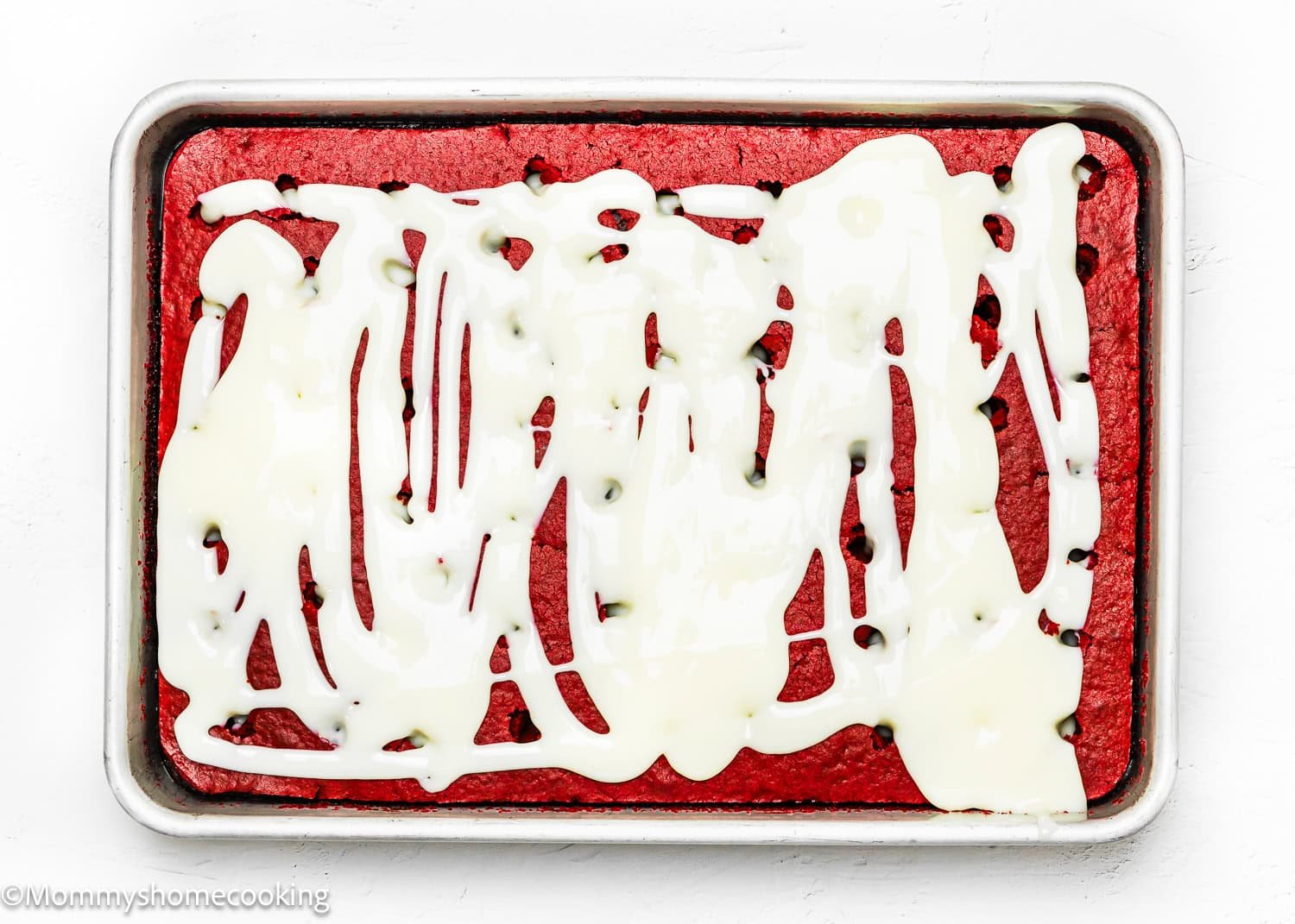 An eggless red velvet poke cake with white icing drizzled on top, set in a metal baking pan, offers a delightful twist on the classic dessert.