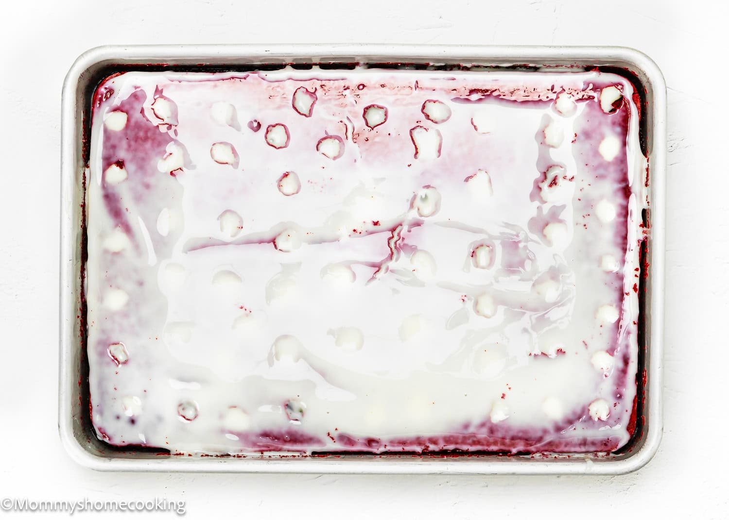 An eggless red velvet brownie poke cake sits on a rectangular tray, its pinkish-red surface frosted with white icing, adorned with scattered droplets of sugar.